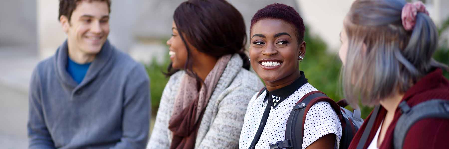 A diverse group of four students sit on a ledge outside with one looking at the camera.